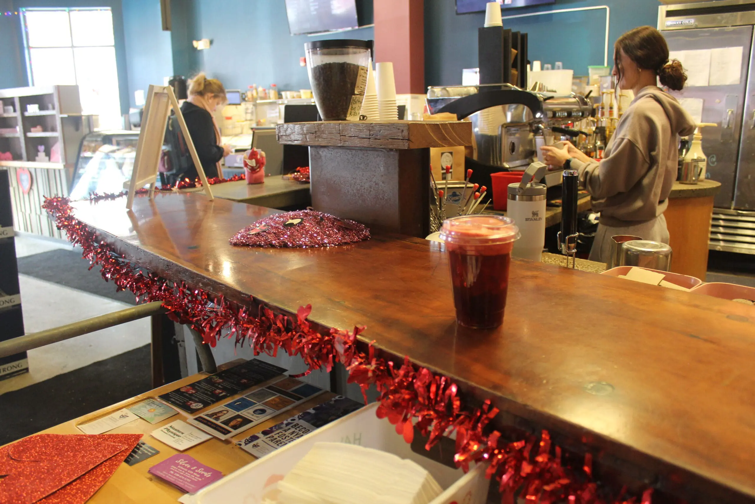 The image shows one of Tree City's baristas making drinks and coffees at an espresso machine. In the distance, a customer can be seen waiting for their coffee at a wooden counter.