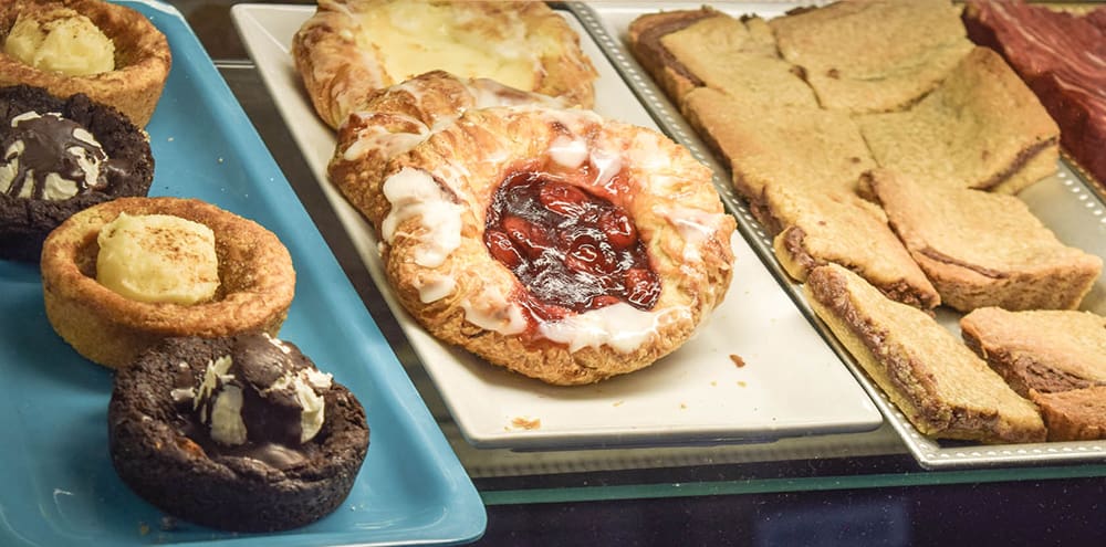 Three trays featuring a variety of pastries. The left tray is blue, and features two brownie bowls with buttercream frosting and hot fudge, and two cinnamon roll bowls with cinnamon frosting. The middle tray is white and features a cherry danish and two cheese danishes. The right tray is gray and is fully stocked with Busy B brownie cookie bars.