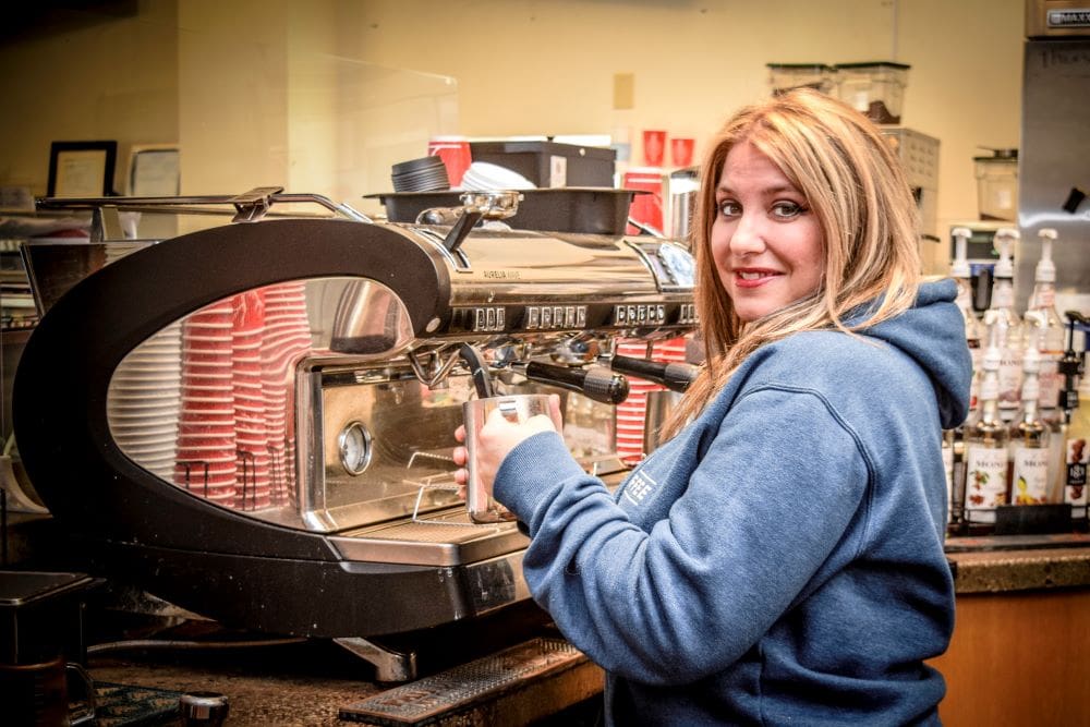 The image features Rebekah, Tree City's Owner, preparing espresso at an espresso machine. Rebekah is holding a metal mug, has long blonde hair, hazel eyes, red lipstick, and wears a blue Tree City pullover sweatshirt.