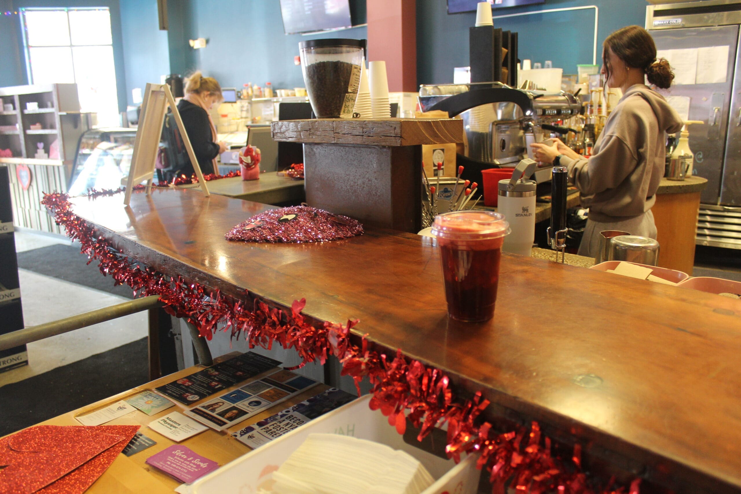 The image shows one of Tree City's baristas making drinks and coffees at an espresso machine. In the distance, a customer can be seen waiting for their coffee at a wooden counter.